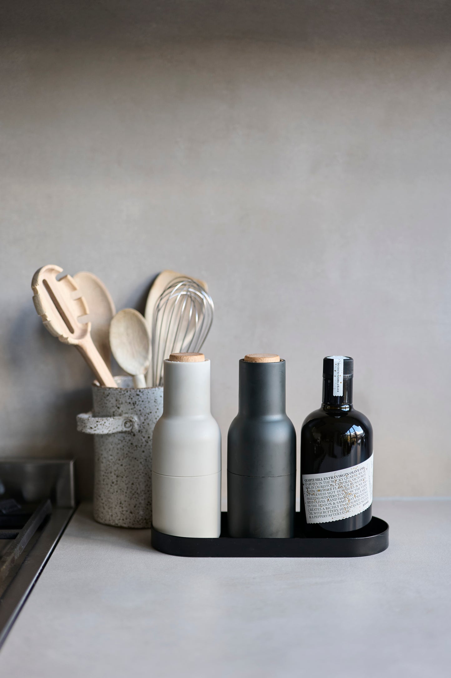 A Bottle of Quartz Hill High Polyphenol Olive Oil on a kitchen bench next to Joseph Joseph salt & pepper shakers, and a ceramic pot filled with wooden cooking utensils