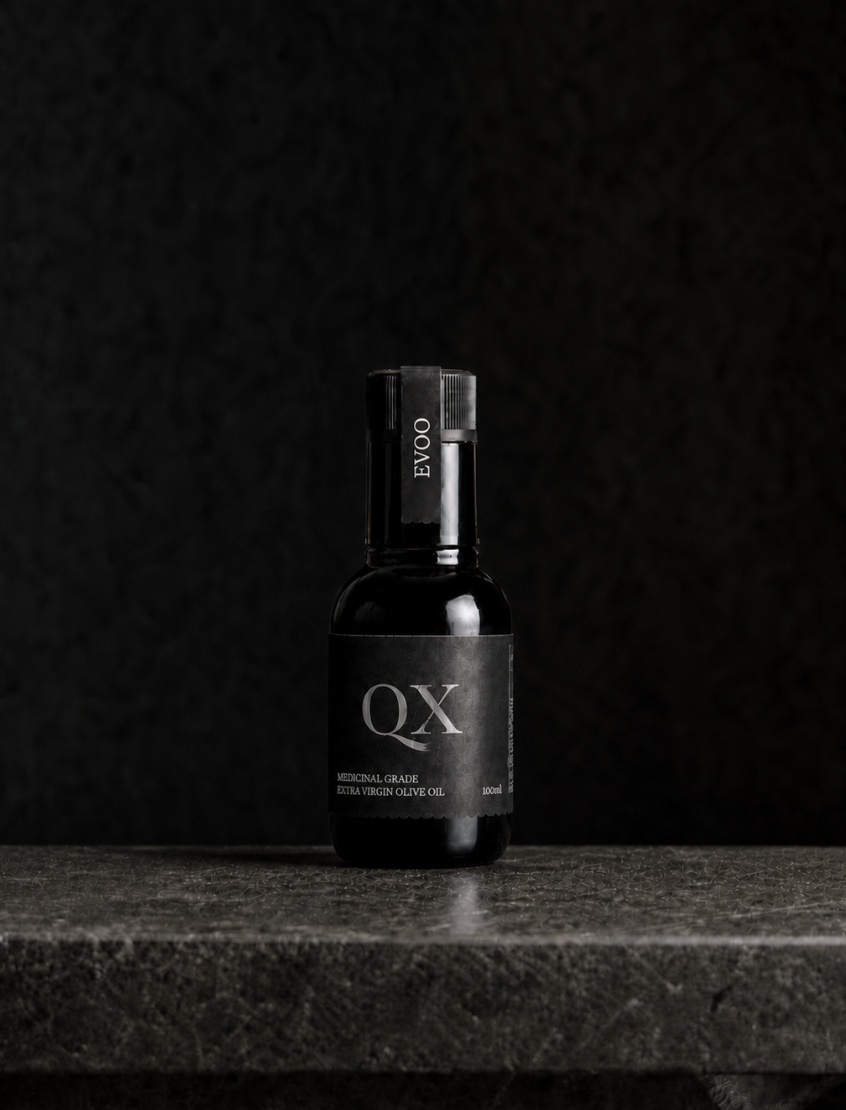 A small black bottle of Australian QX Medicinal Grade Olive Oil with a black cap, standing on a dark stone surface against a dark textured background.