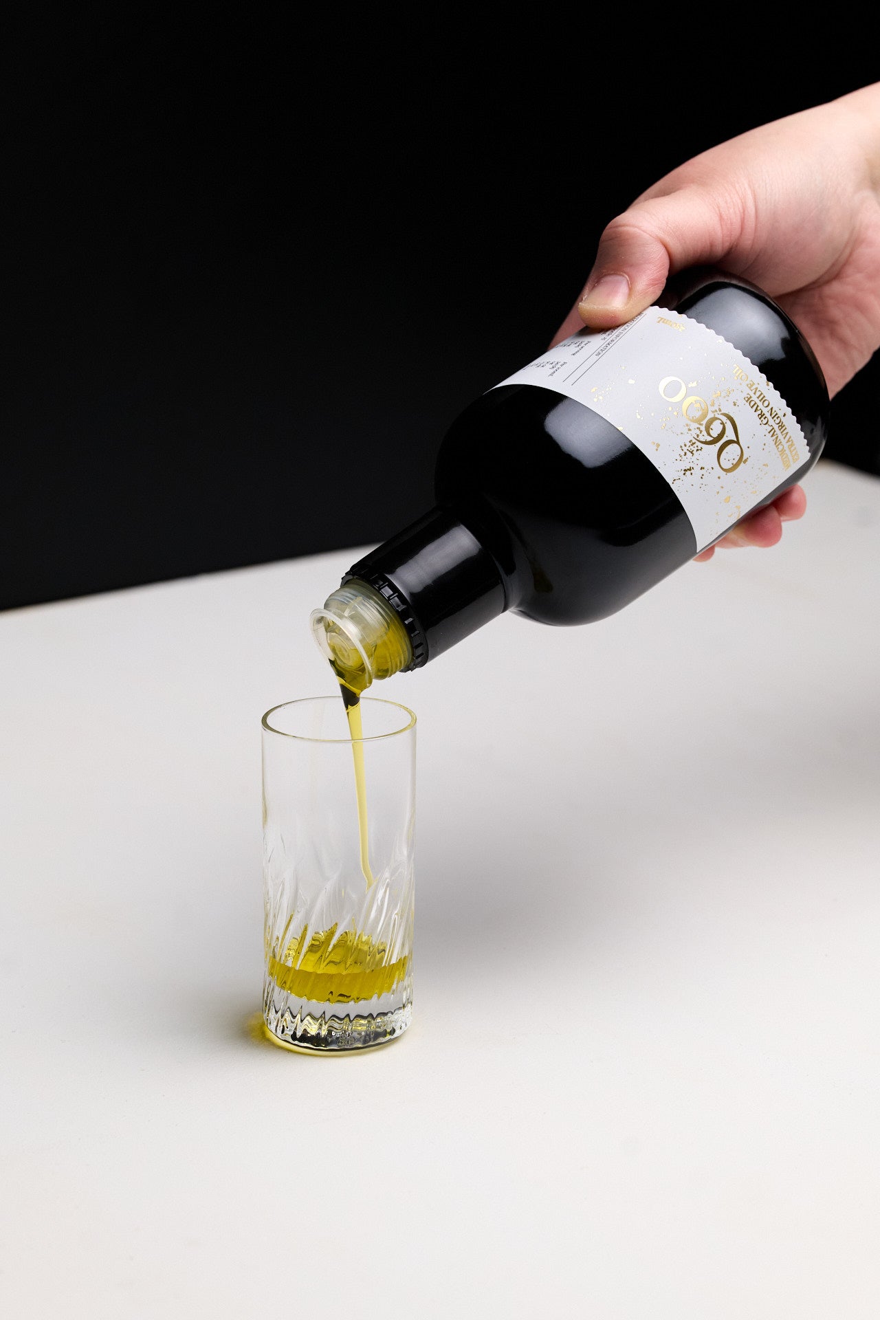 Daily shot of olive oil being displayed with a hand high polyphenol olive oil from a bottle into a glass on a white surface with a black background.