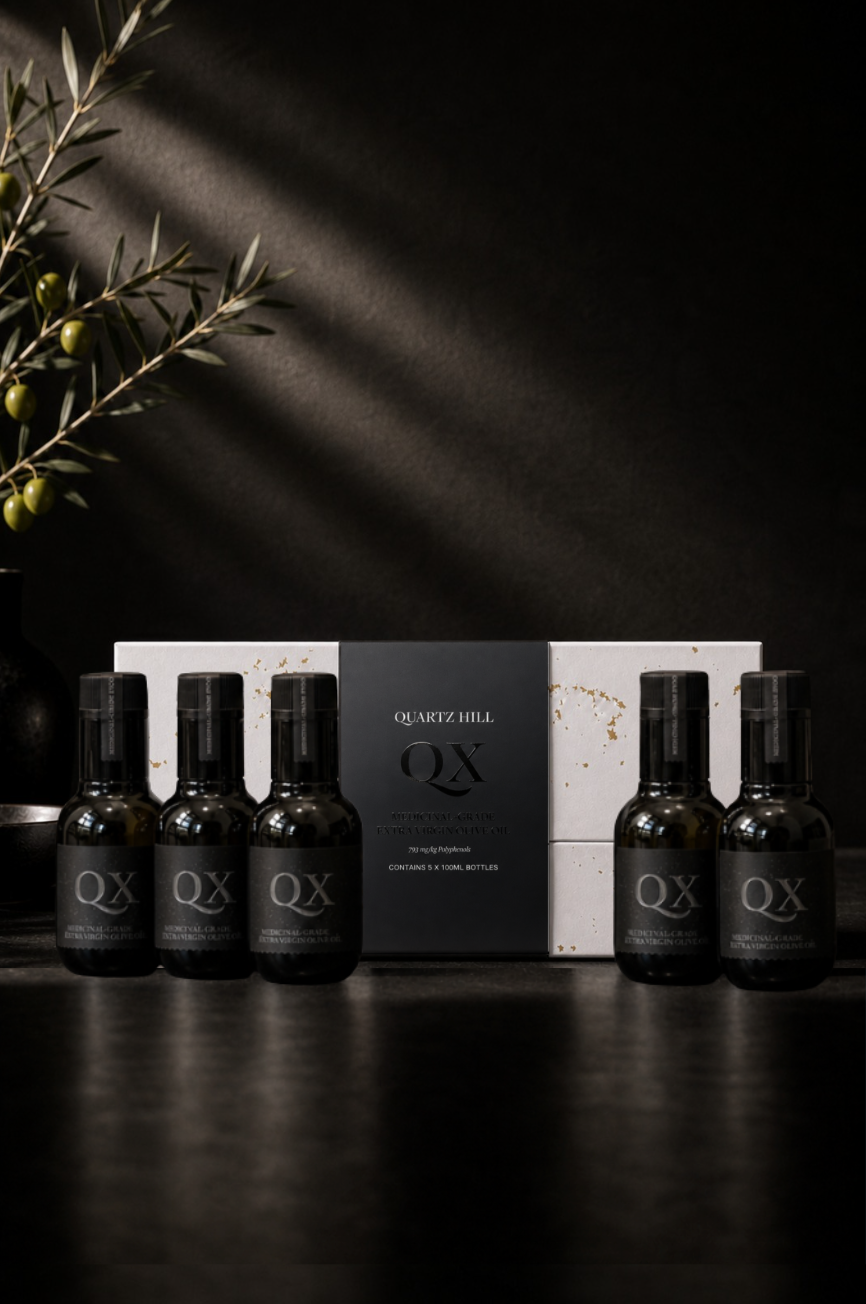 Quartz Hill QX 30-day high-polyphenol olive oil challenge pack featuring five 100ml medicinal-grade dark glass bottles arranged in front of a luxury white and gold gift box under moody lighting next to an olive branch.