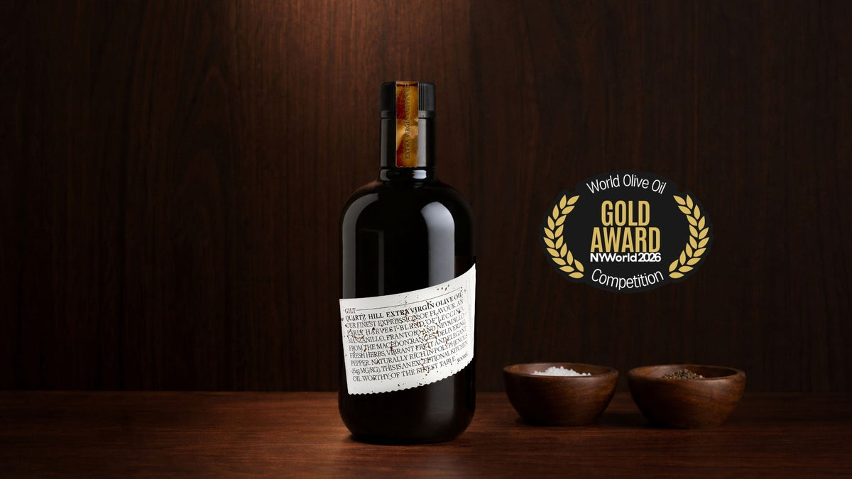 Quartz Hill Gilt extra virgin high-polyphenol olive oil on a wood bench showing the award for gold at the NYIOOC World Olive Oil Competition next to the black glass bottle.