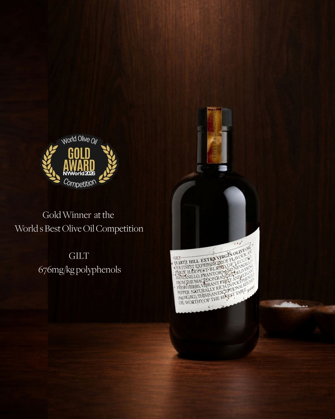 Quartz Hill Gilt extra virgin olive oil on a wood bench showing the award for gold at the NYIOOC World Olive Oil Competition next to the black glass bottle.