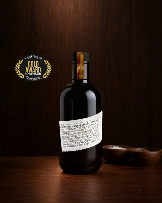 Quartz Hill Gilt extra virgin olive oil on a wood bench showing the award for gold at the NYIOOC World Olive Oil Competition next to the black glass bottle.