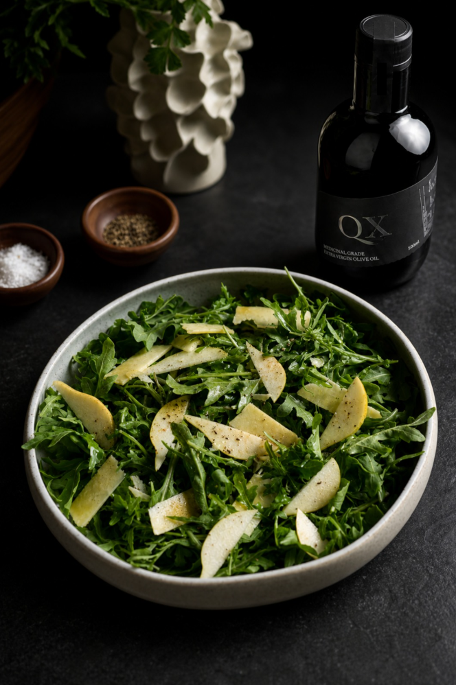 A bowl of fresh arugula salad topped with sliced pears and a drizzle of oil, styled on a dark tabletop with a bottle of QX olive oil, small bowls of seasoning, and a decorative vase in the background.