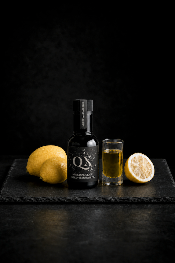A single 100ml dark glass bottle of QX Medicinal-Grade Extra Virgin Olive Oil next to a filled glass shot glass, two whole lemons, and a halved lemon on a textured black slate board against a dark background for olive oil lemon juice wellness shots.